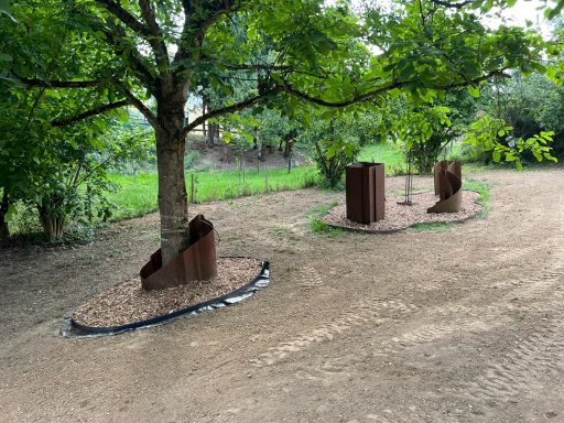 Skulpturen aus Cortenstahl unter einem Baum auf einem leicht unebenen, sandigen Boden.