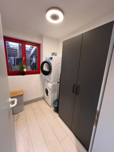 Heller Raum mit Waschmaschine, Trockner und einem hohen Schrank. Rote Fensterrahmen.
