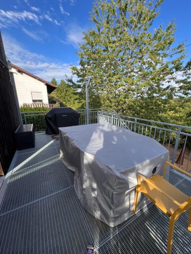 Balkon mit einem Tisch, einer Bank und Blick auf Bäume und den Himmel.