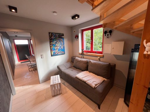 Modern Wohnraum mit grauem Sofa, Fenster mit rotem Rahmen und Holztreppe.